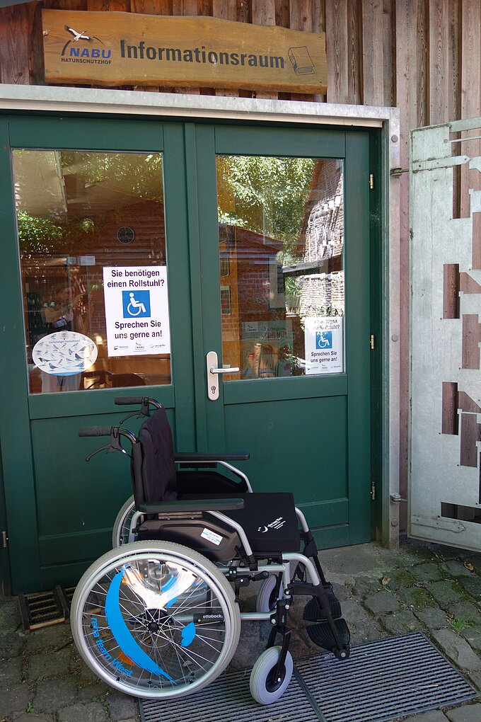 Der geländegängige Rollstuhl vor der Eingangstür des Seminarraums. An der Tür informeiert ein Schild über die Verfügbarkeit des Rollstuhls.